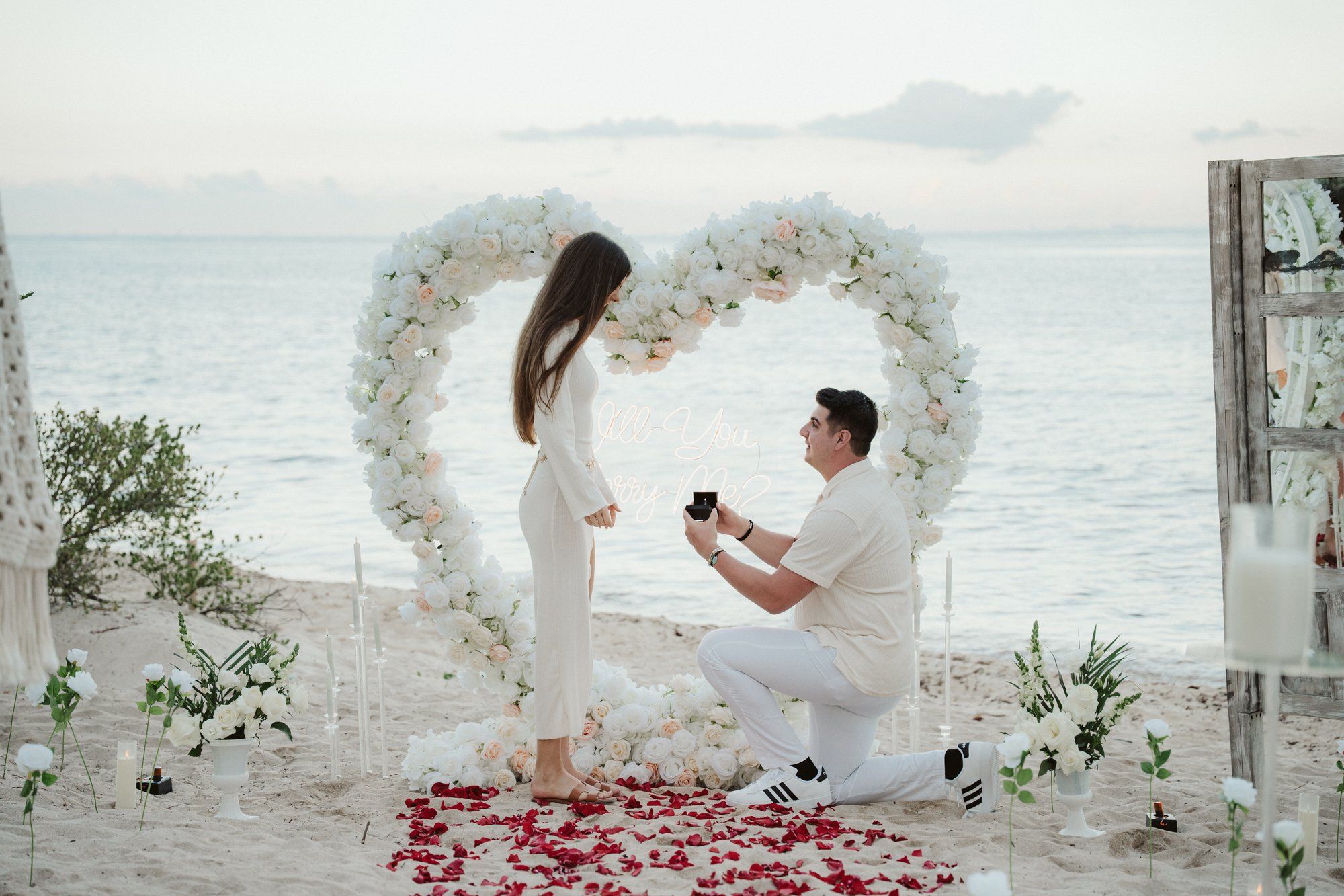 Dramatic sunset proposal in Playa del Carmen with a giant rose heart arch positioned perfectly against the horizon and crashing waves.