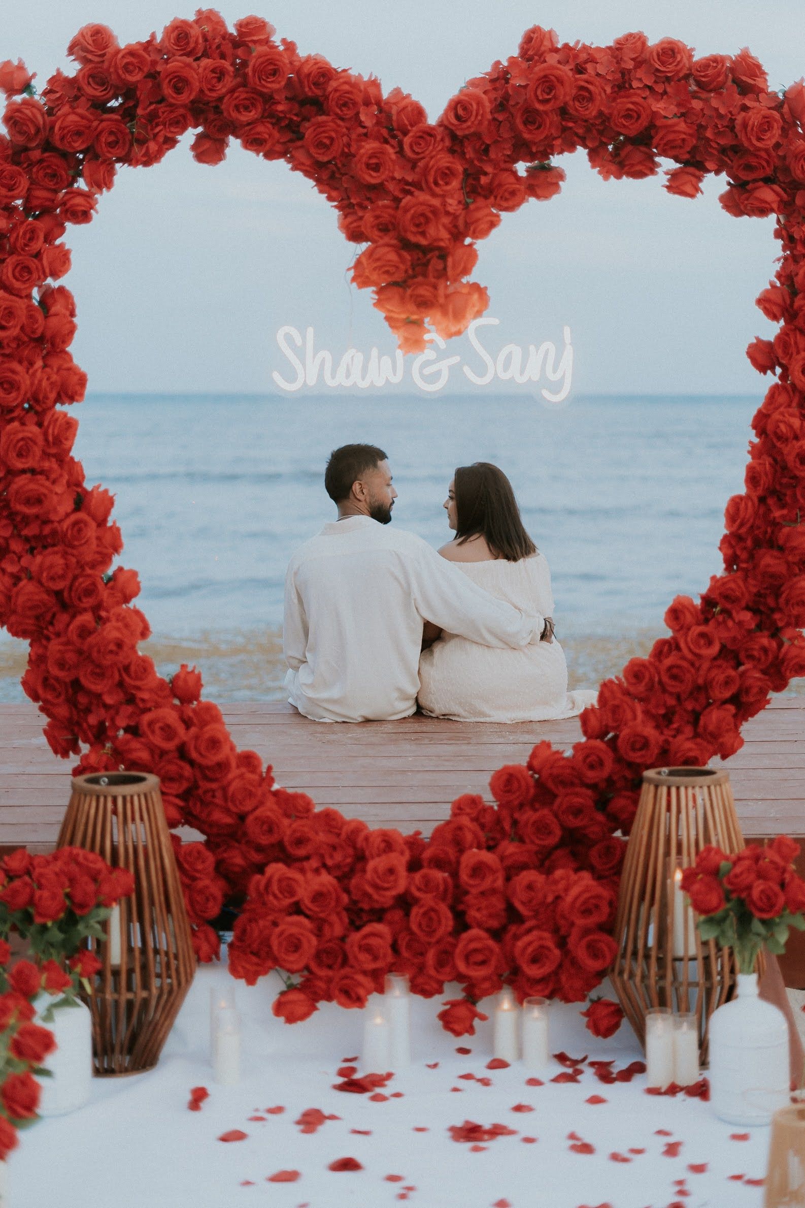 Luxury beach proposal in Tulum featuring a long pathway of red rose petals leading to a floral heart arch and 'Will You Marry Me?' neon sign at twilight.