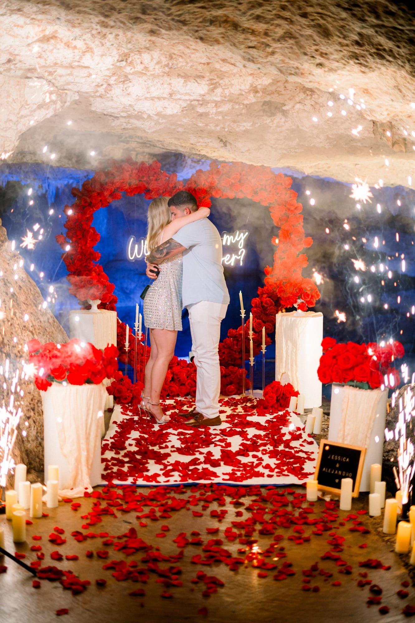 Pareja abrazándose bajo chisperos fríos en una cueva de cenote en Riviera Maya, enmarcada por un arco de corazón de rosas rojas, velas y pétalos.