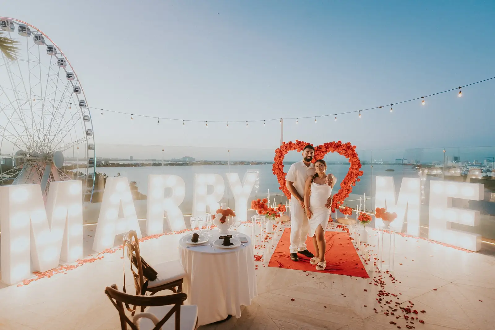 Celebración de propuesta de matrimonio en una azotea con flores y letras iluminadas