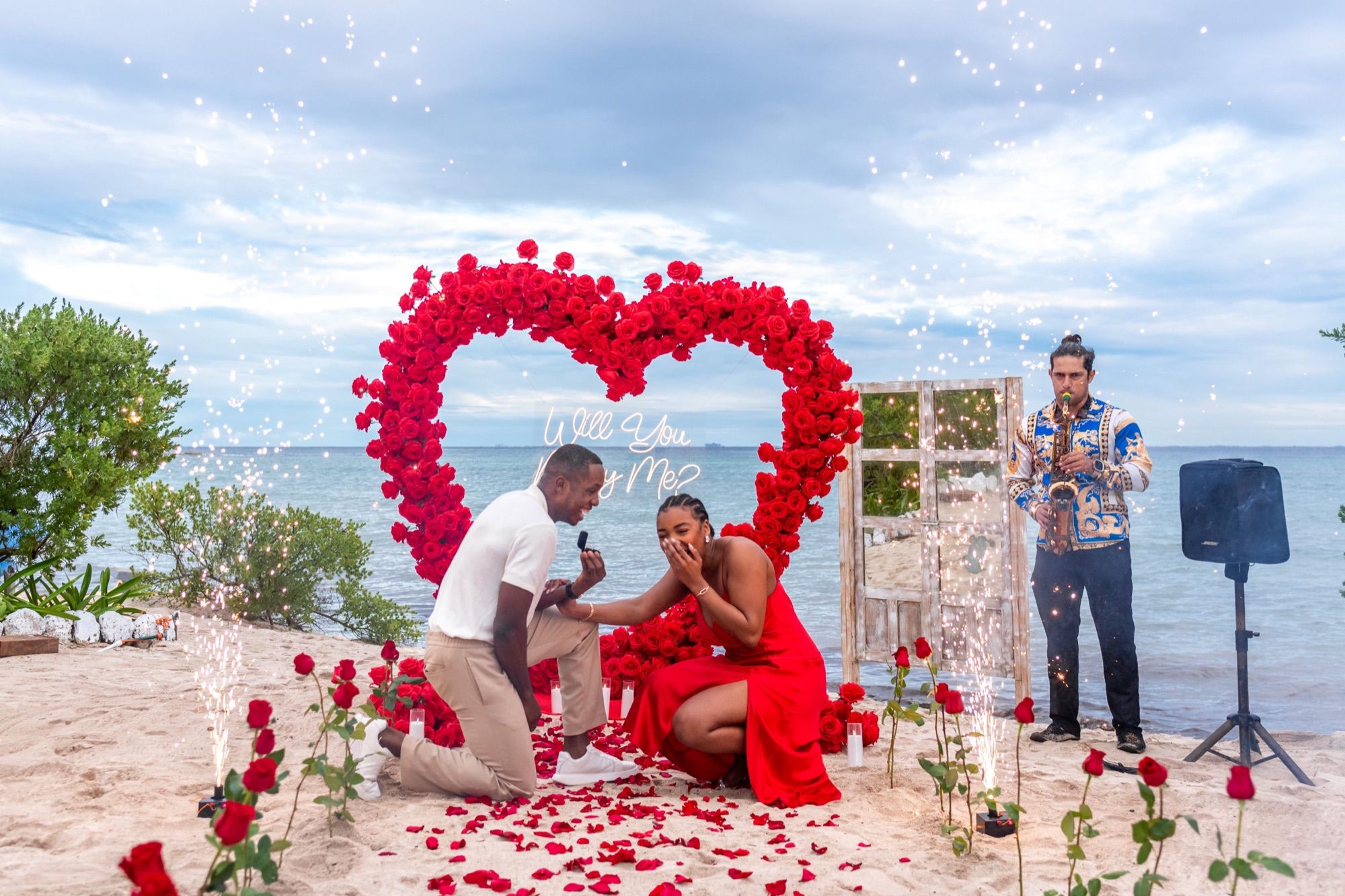 Momento de propuesta Signature en la playa con arco de corazón de rosas rojas, saxofonista en vivo y chisperos fríos